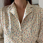 70s Delightful feminine shaped blouse in a lightweight fabric with floral printed pattern. French feminine and romantic style piece.   •	Long sleeves •	Shirt collar •	Features a peplum hem •	Accented by a row of covered buttons down the front •	Adorned with delicate smocking detail at front •	Half-lined 